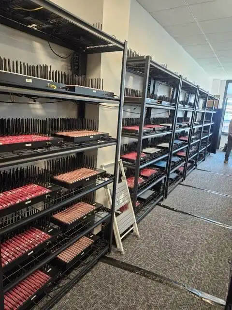 Zdjęcie z aresztu pokazujące zabezpieczone urządzenia z farmy SIM: regały pełne smartfonów i modułów GSM używanych do nielegalnej rejestracji kont i ataków phishingowych na dużą skalę